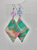 A pair of handmade dangle earrings with teal and pink marbled resin triangles, gold foil accents, and iridescent blue-purple glass beads on silver fishhook wires, displayed on a clear stand against a light gray background.