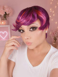 Woman with vibrant pink and purple hair in a stylish pose against a pink background with decorative elements.