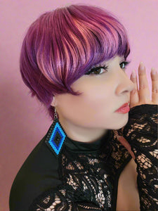 Person with vibrant purple hair wearing a black lace top and blue earrings.