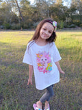Young girl standing in a grassy field wearing a white t-shirt with a colorful design.
