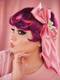 Woman with pink hair styled in pigtails with a large bow, wearing a pink sweater against a pink background.