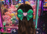 Large green pixel art bow hair clip with a pink heart center styled in a half-up hairstyle in front of an arcade claw machine